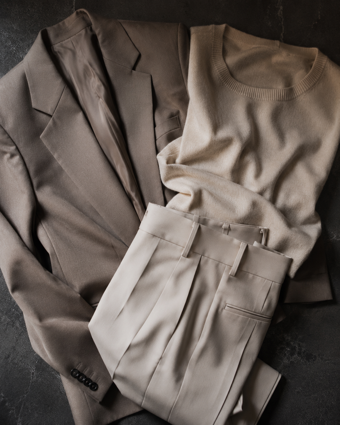 A folded taupe blazer layered over a light beige crewneck sweater and matching pleated trousers, arranged in a flat lay on a dark textured surface.