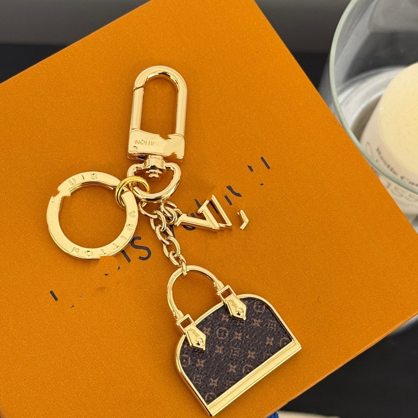 A Louis Vuitton monogrammed gold-tone keychain featuring a miniature handbag charm, displayed on an orange Louis Vuitton box.