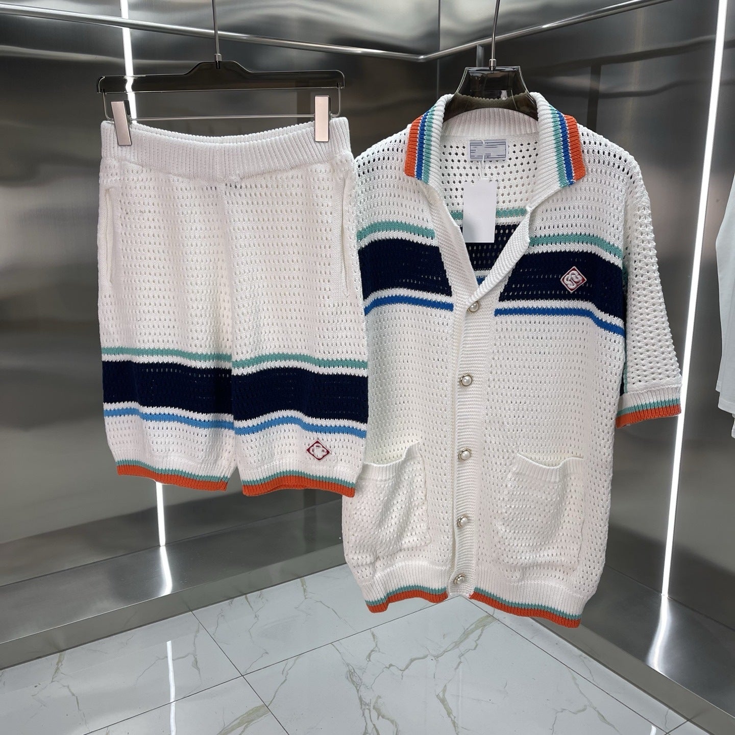 A matching knit two-piece set hanging on black hangers against a metallic wall. The set includes white knit shorts and a matching open-front cardigan with navy, teal, and orange stripes.