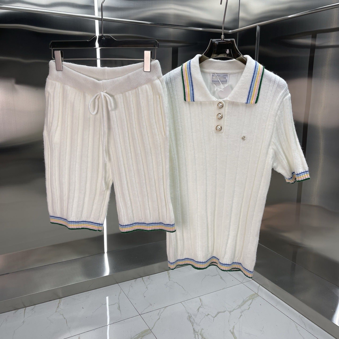 A matching two-piece knit set consisting of white ribbed shorts with a drawstring waist and a short-sleeve polo shirt with striped trim, hanging on black hangers against a metallic wall.