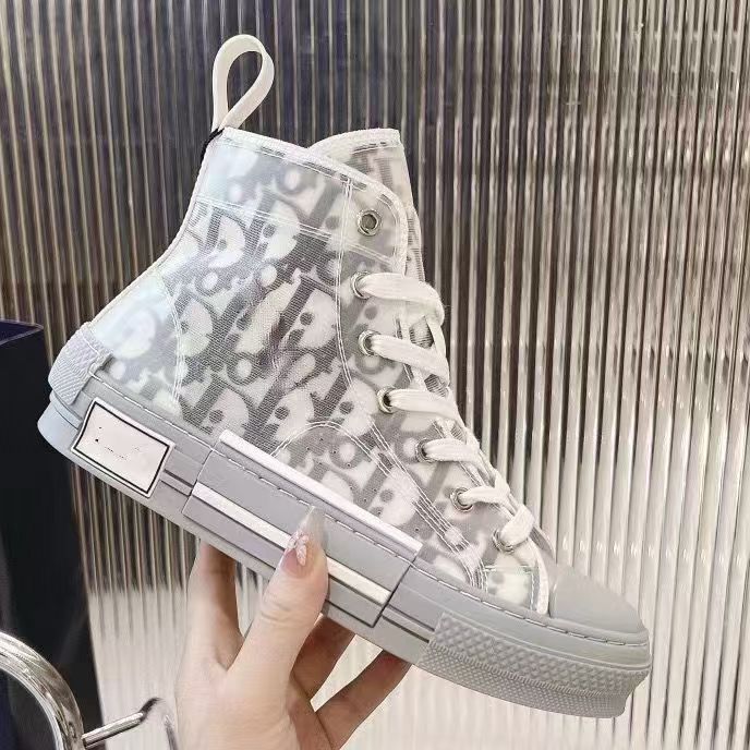 A hand holds a high-top sneaker featuring a white and gray Dior monogram canvas upper, white laces, and a gray rubber sole, set against a metallic ribbed background.