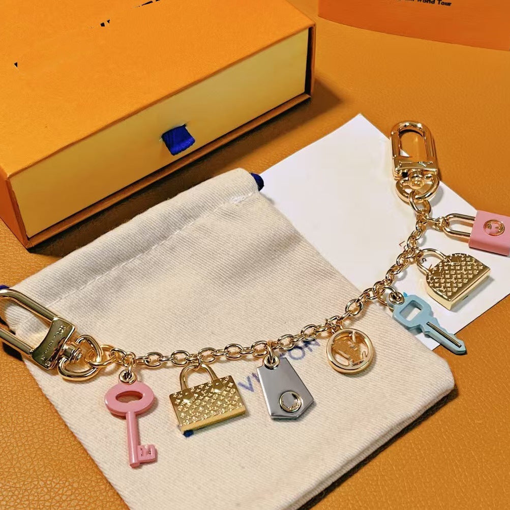 A gold-tone keychain charm bracelet with multiple decorative charms, including a pink key, a pink padlock, a gold padlock, a silver padlock, a light blue key, and a gold circular charm, displayed on a white fabric pouch with an orange box in the background.