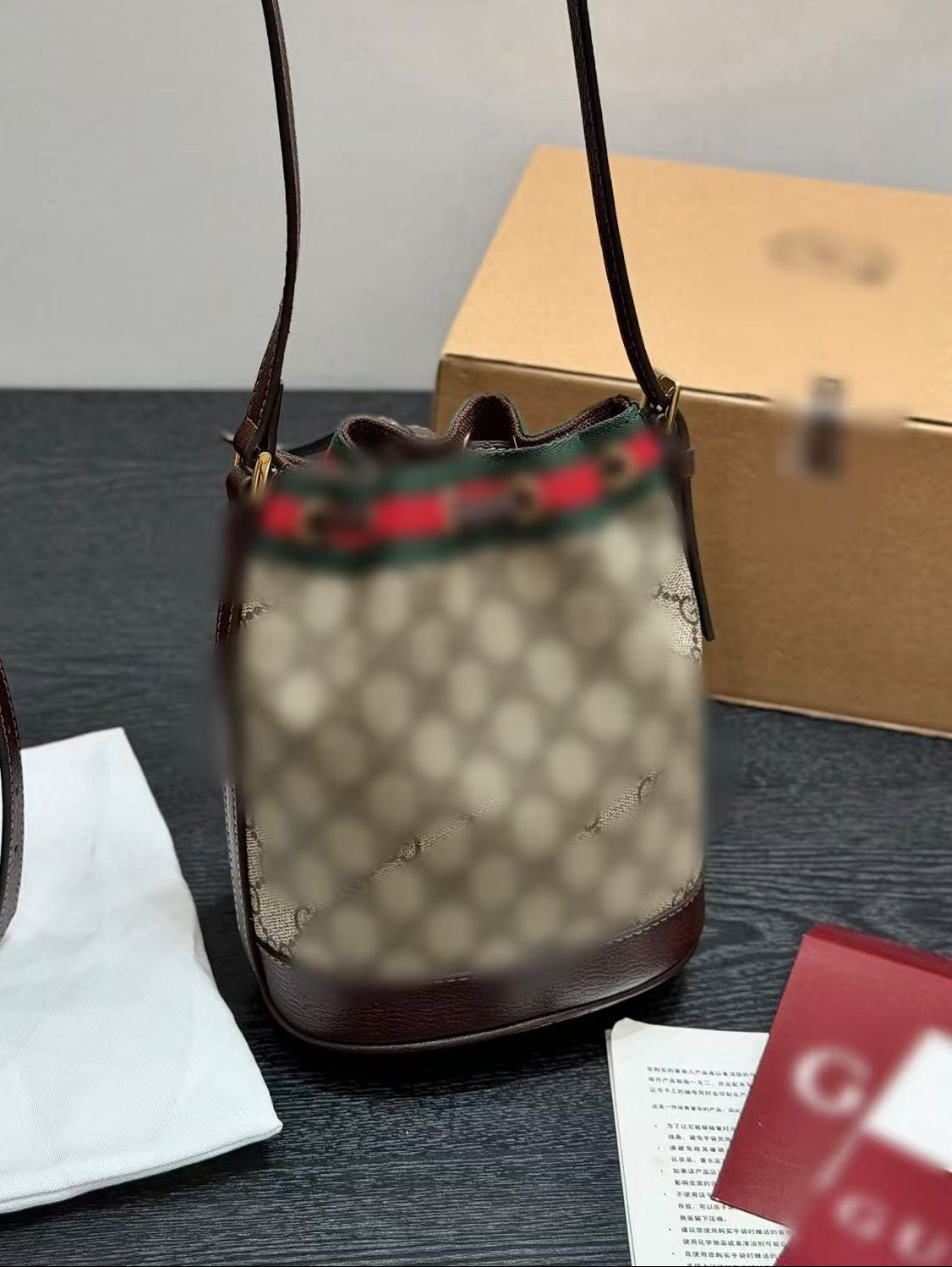 A Gucci bucket bag with beige GG monogram canvas, dark brown leather trim, and the signature red-green-green web stripe detail at the top, shown on a tabletop with its box and dust bag nearby.