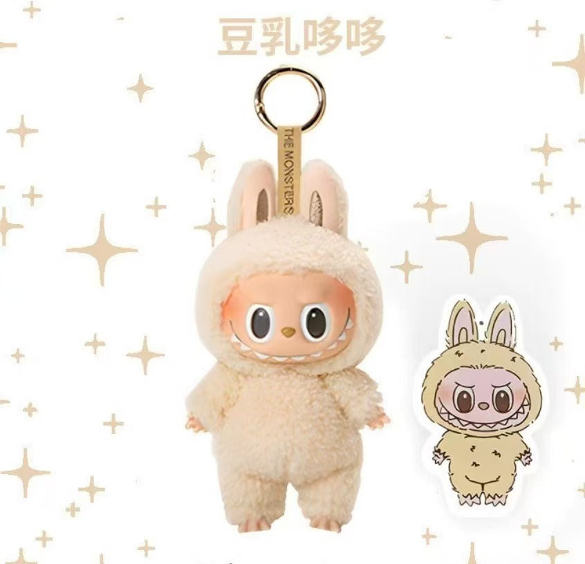 A plush character keychain in a cream-colored fuzzy bunny suit with a gold key ring, shown alongside a small die-cut sticker of the same character on a white background with gold star accents.