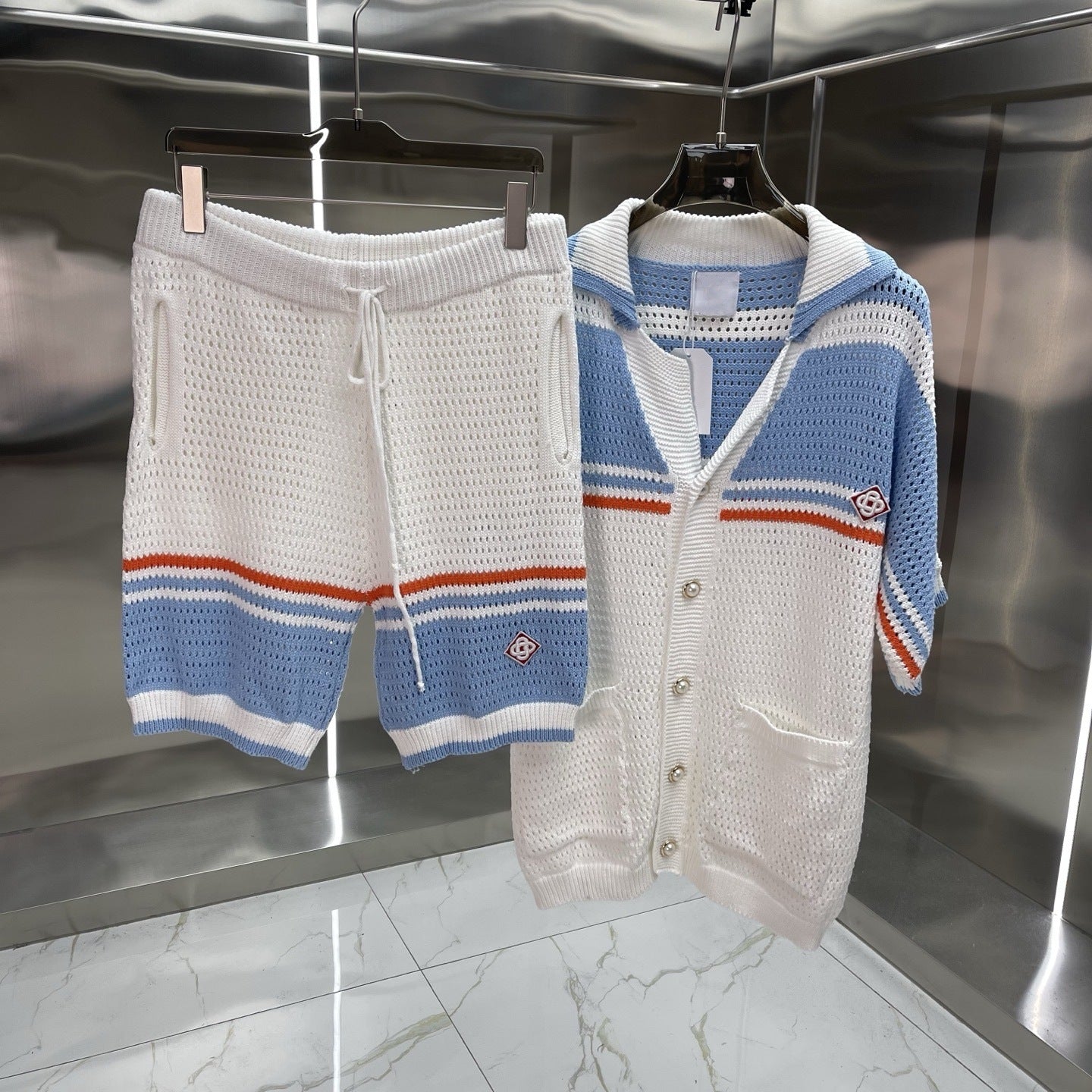 A knit two-piece clothing set hanging on black hangers against a metallic wall. The set includes white knit shorts with blue and orange stripes at the hem and a matching white knit cardigan with blue horizontal stripes and orange accents.