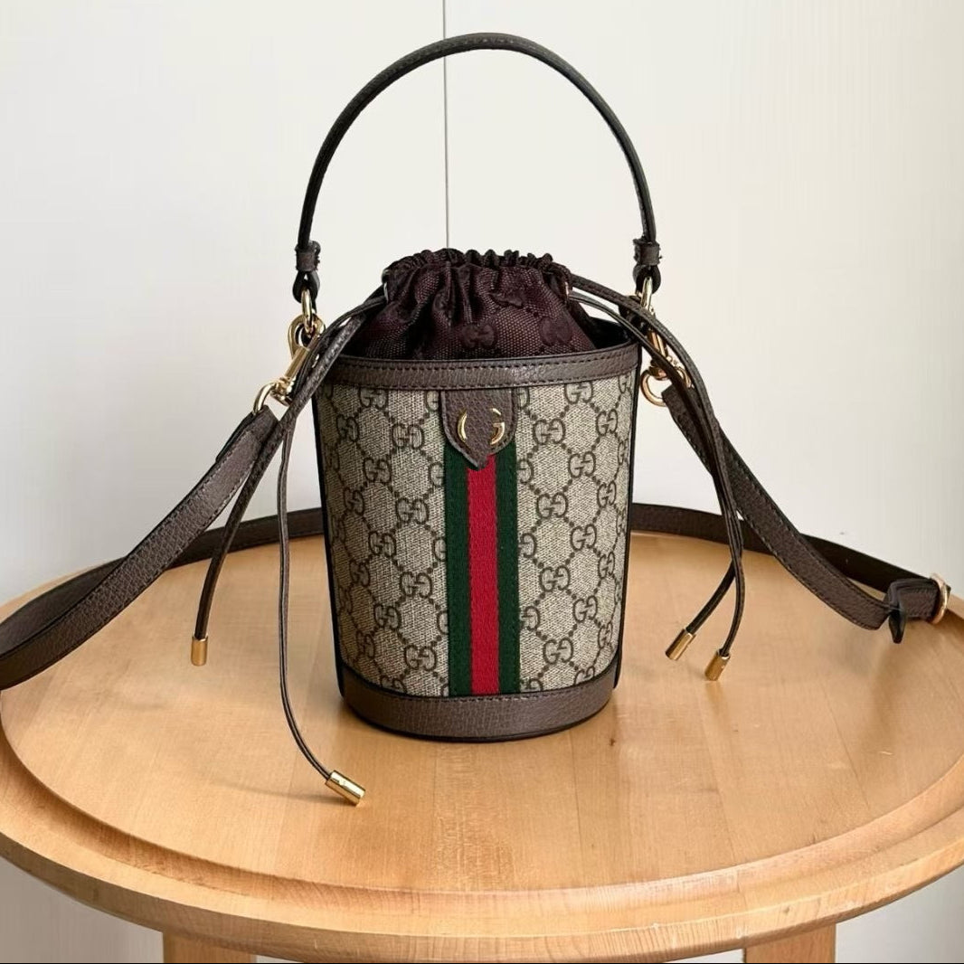 A Gucci GG Supreme bucket bag with a dark brown leather handle and shoulder strap, featuring the classic beige and brown monogram pattern and green and red web stripe. The bag is displayed on a round wooden table against a plain off-white wall, with Chinese text overlays indicating the style number and dimensions.