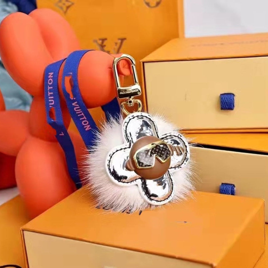 A Louis Vuitton-inspired keychain charm with a white faux-fur pom-pom, a silver-tone flower-shaped clasp, and a blue strap printed with 'LOUIS VUITTON', displayed among orange Louis Vuitton boxes.