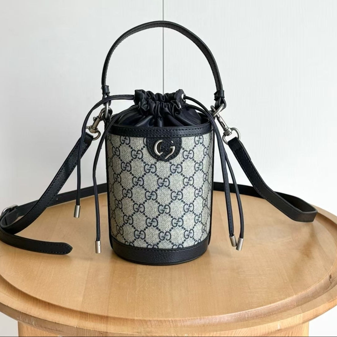 A small cylindrical bucket bag with a black leather top handle, shoulder strap, and trim, featuring a beige and blue repeating monogram pattern on the body. The bag is displayed on a round wooden table against a plain white wall, with Chinese text overlay in the top-left corner showing the style number and dimensions.