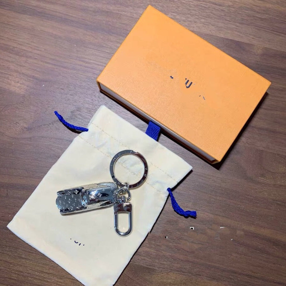 A silver metal keychain with a nail clipper attachment, displayed on a cream-colored drawstring pouch next to an orange box on a dark wood surface.