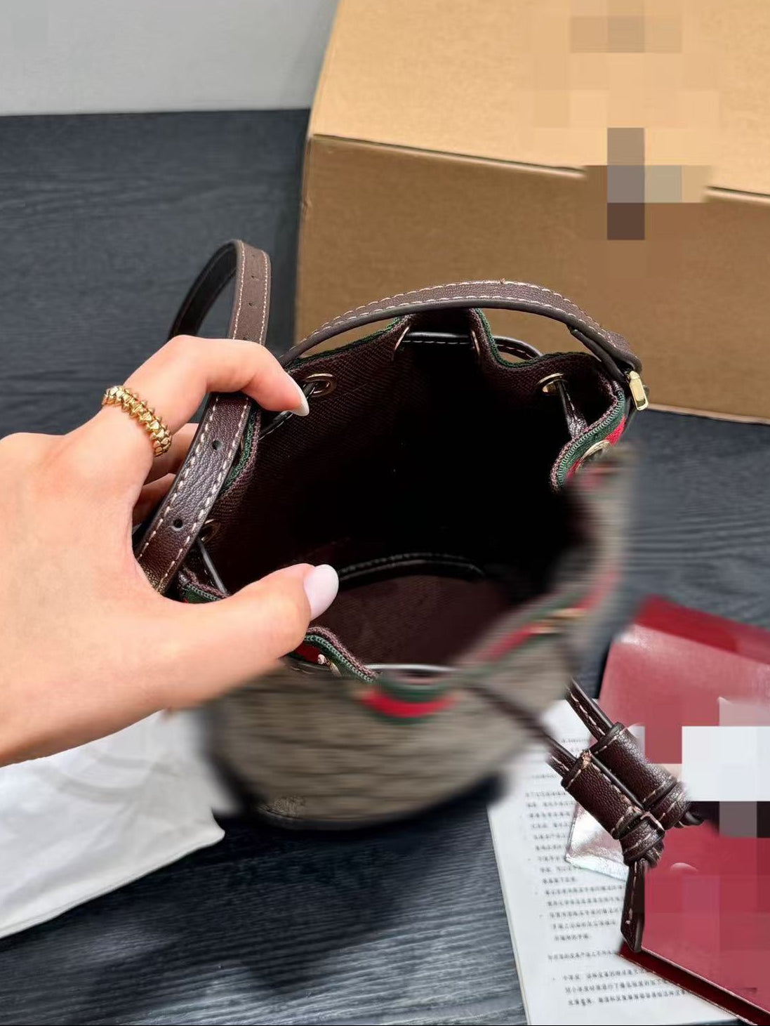 A hand holding the open top of a brown bucket bag with a dark brown interior, shown on a tabletop next to a cardboard box and papers. Chinese text is visible at the top.