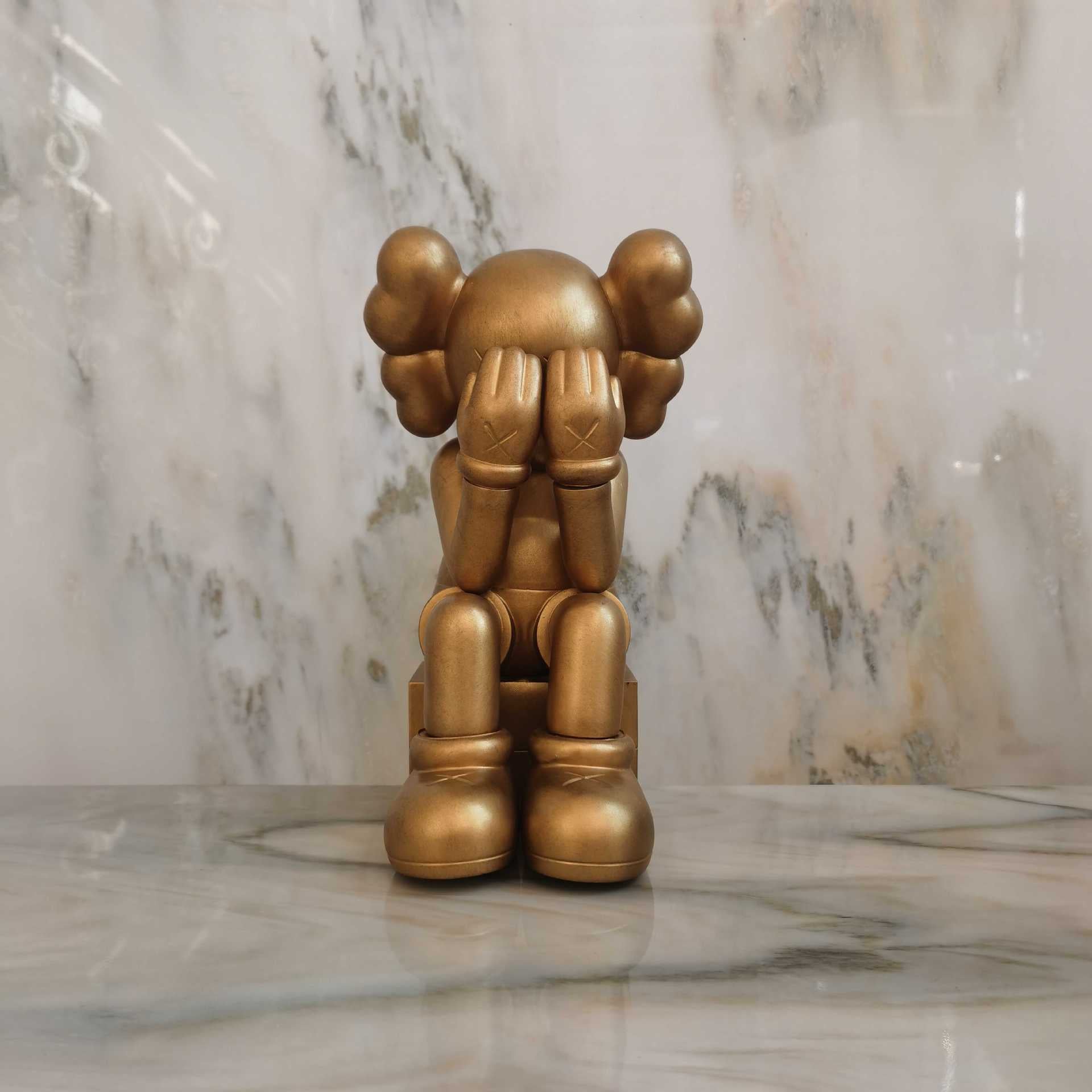 A gold-colored KAWS-style character figurine sitting on a marble surface with a matching marble background.