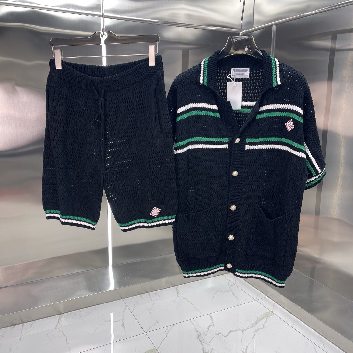 A matching knit lounge set consisting of a short-sleeve button-front cardigan and shorts, both in black with green and white striped accents, hanging on hangers against a metallic silver wall.