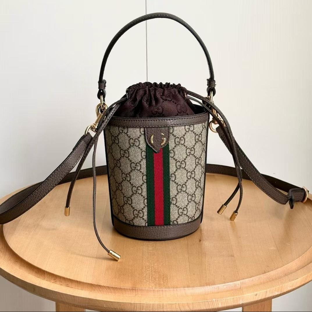 A Gucci GG Supreme bucket bag with a dark brown leather handle and shoulder strap, featuring the classic beige and brown monogram pattern and green and red web stripe. The bag is displayed on a round wooden table against a plain off-white wall, with Chinese text overlays indicating the style number and dimensions.