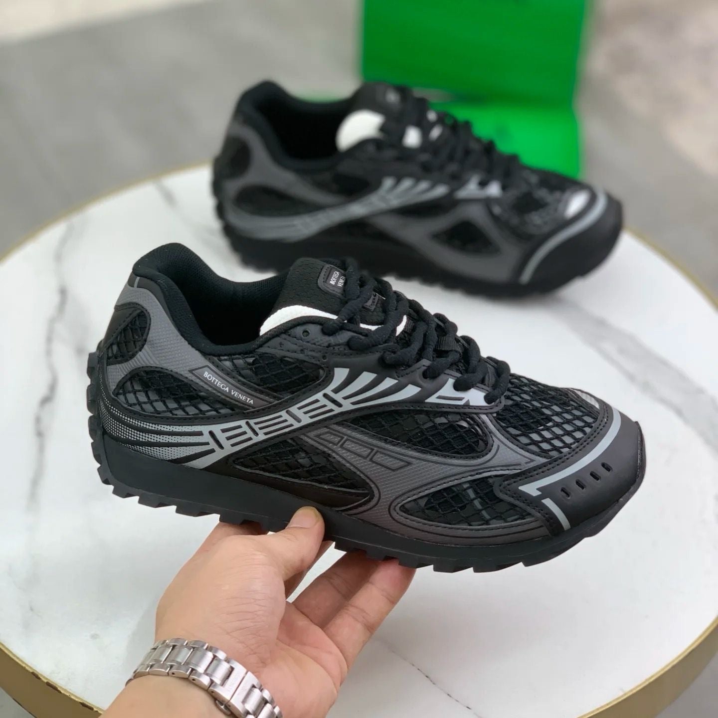 Black and gray athletic shoes held by a hand with a silver watch on a white surface.