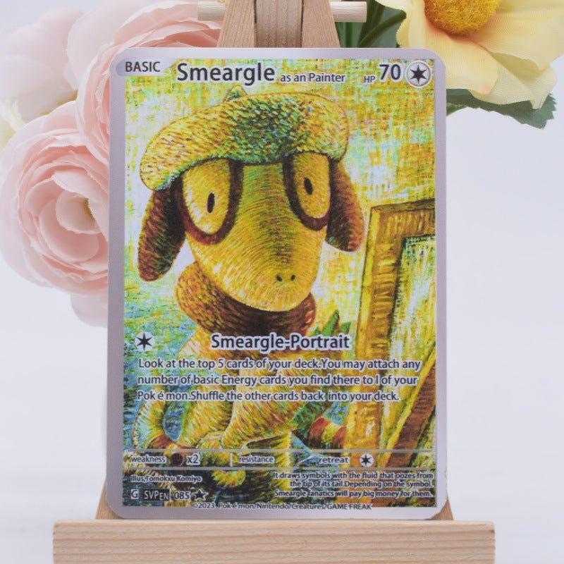 A Pokémon Smearle trading card displayed upright on a small wooden stand, with soft pink and yellow flowers blurred in the background.