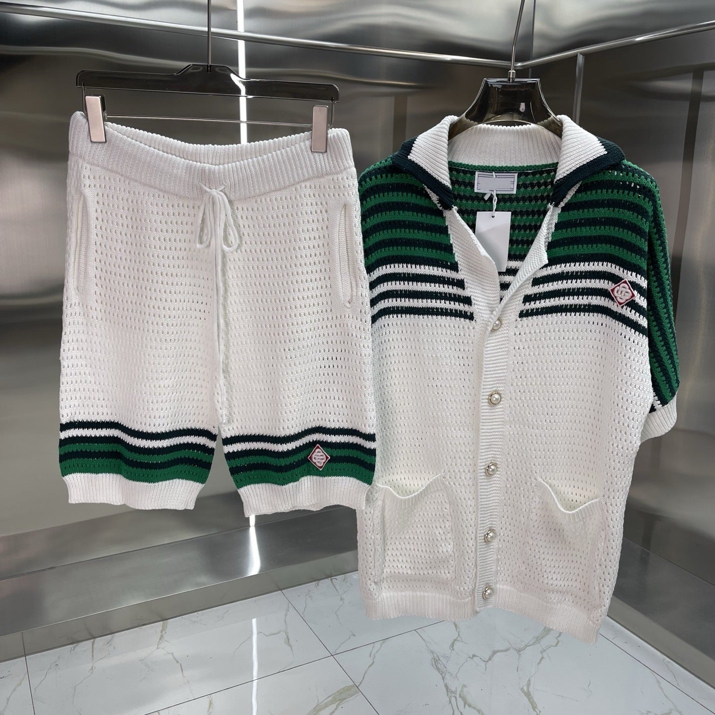 A knit two-piece outfit displayed on hangers against a metallic wall, featuring white textured shorts with green and navy stripes and a matching white cardigan with green and navy horizontal stripes.