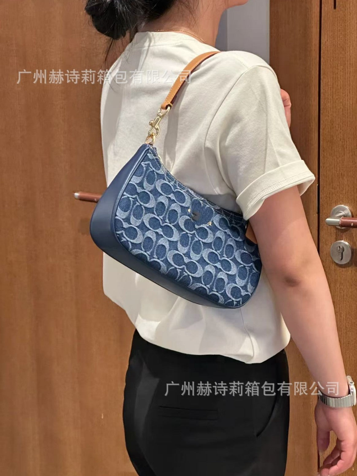 Person wearing a blue handbag with a brand logo, standing in front of a wooden door.
