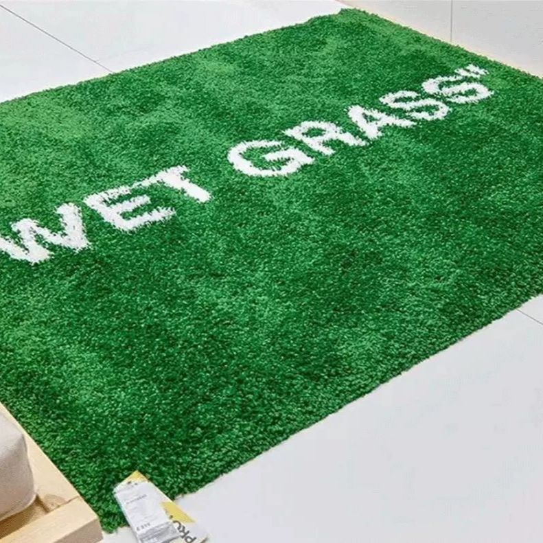 A green artificial turf-style rug with white fuzzy text reading 'WET GRASS' across the surface, photographed on a light floor.