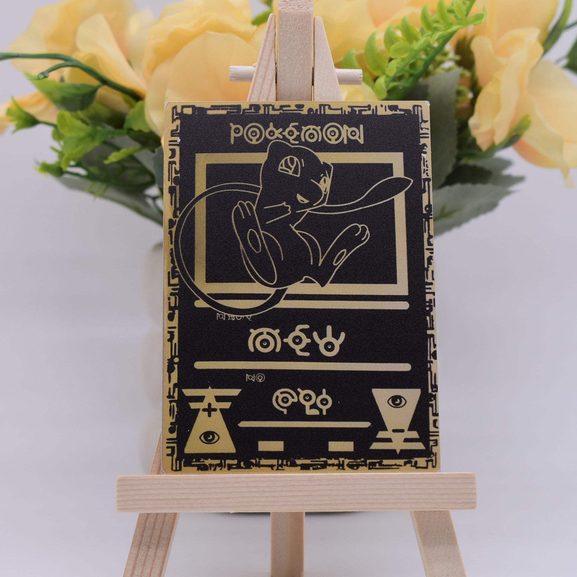 A small black rectangular card or plaque with gold line art and text, displayed on a miniature wooden easel in front of yellow flowers.