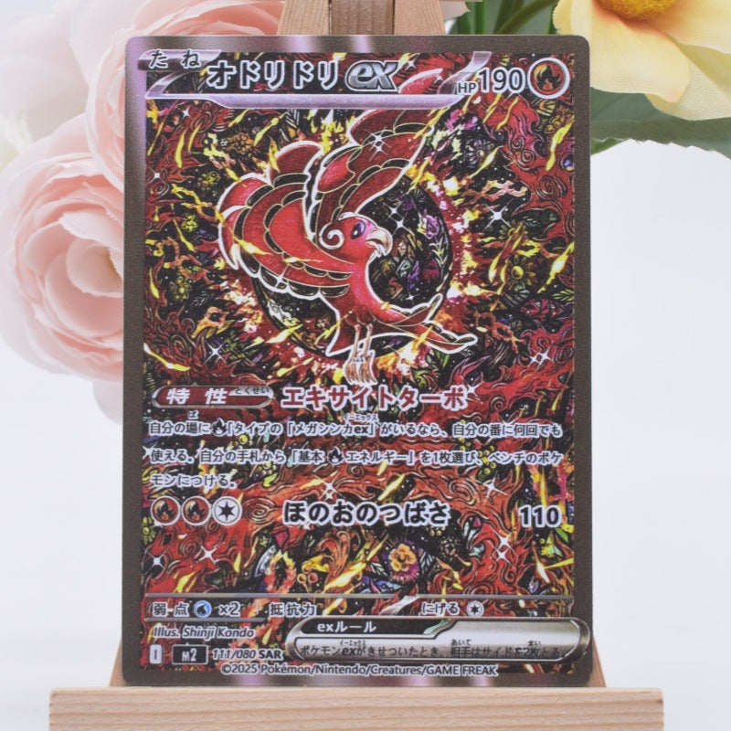 A Japanese Pokémon trading card featuring the ex. Cinderace card, displayed on a small wooden stand with soft pink and yellow flowers in the background.