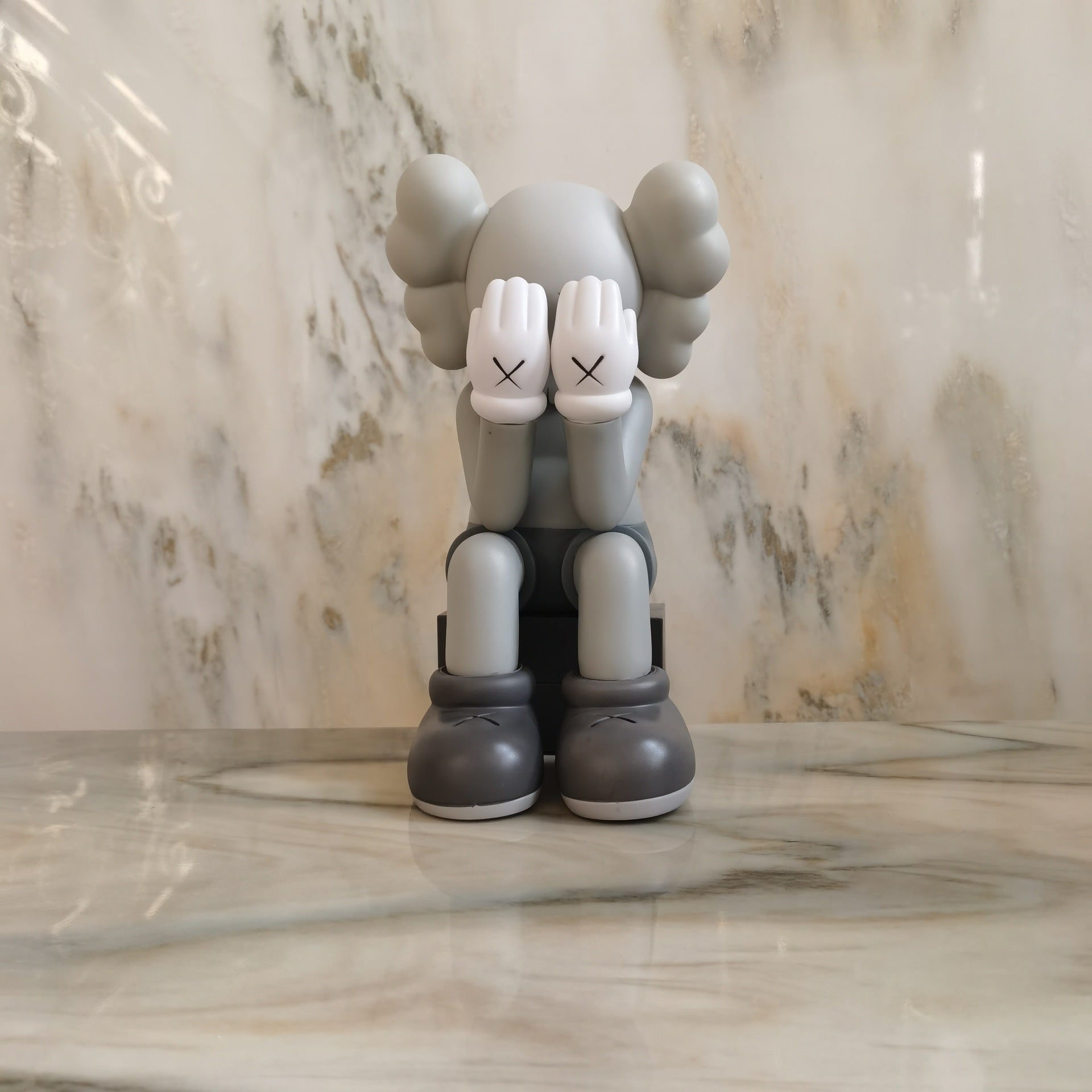 A gray and white KAWS character figurine sitting on a marble surface against a marble background.
