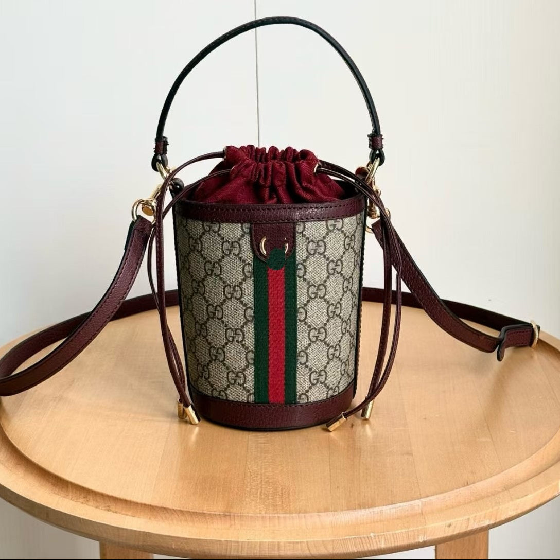 A Gucci bucket bag with the classic GG monogram pattern, dark brown leather trim, a green and red web stripe, and a dark red drawstring top. The bag is displayed on a round wooden table against a plain white wall, with Chinese text in the upper left indicating the style number and dimensions.