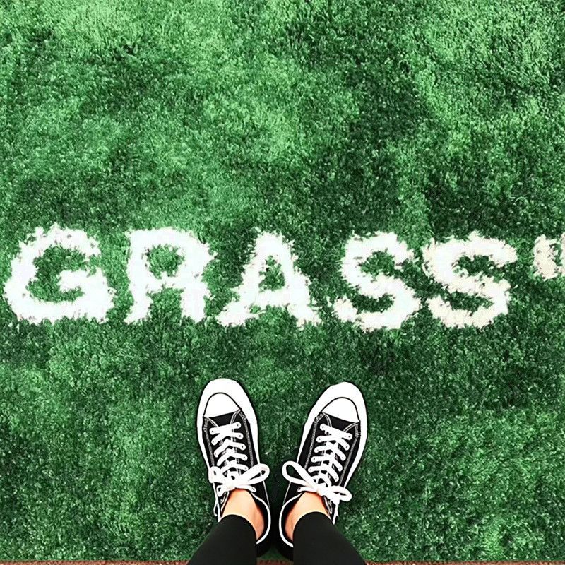 A top-down view of a person's feet in black and white canvas sneakers standing on a green artificial turf field with the word 'GRASS' spelled out in white letters on the turf.