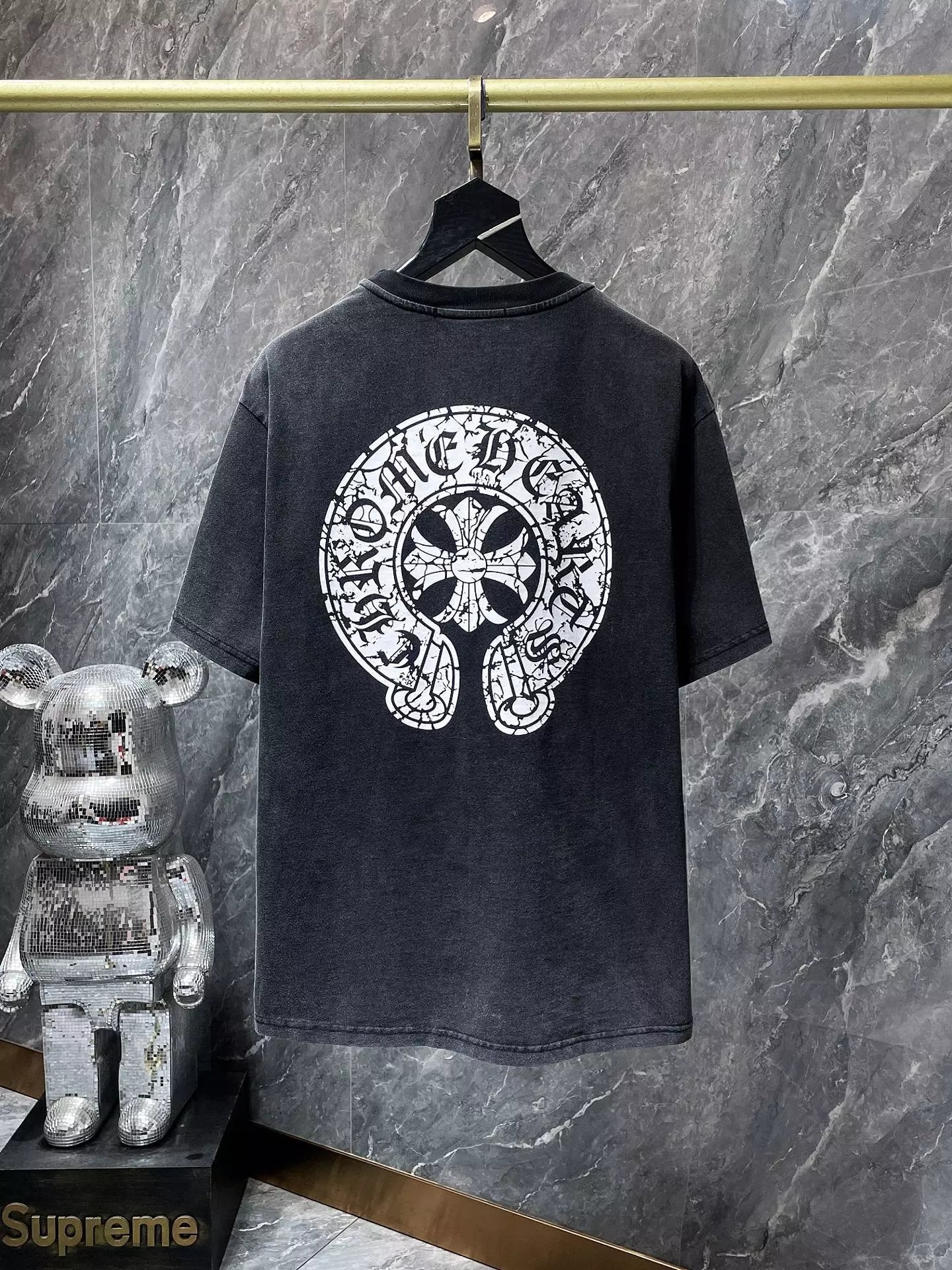 Black t-shirt with a white logo on a hanger against a marble wall, with a Supreme toy in the foreground.