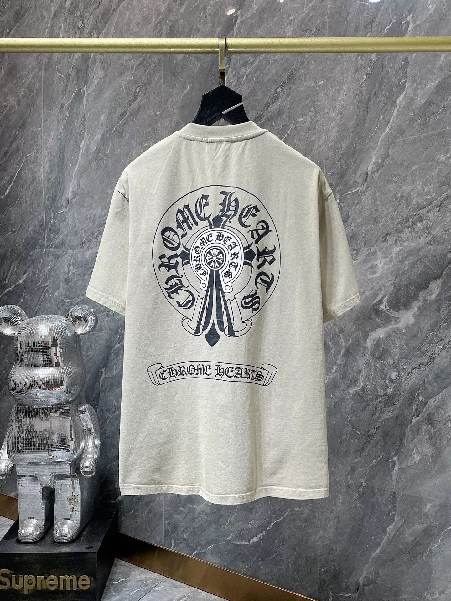 Beige t-shirt with black graphic design on a hanger against a gray marble wall.