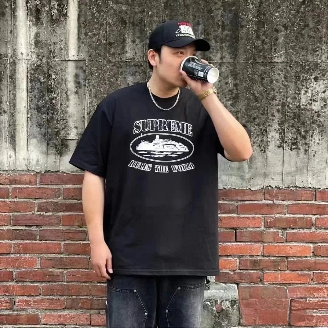 Person wearing a black 'Supreme' t-shirt and cap, drinking from a can against a brick wall.