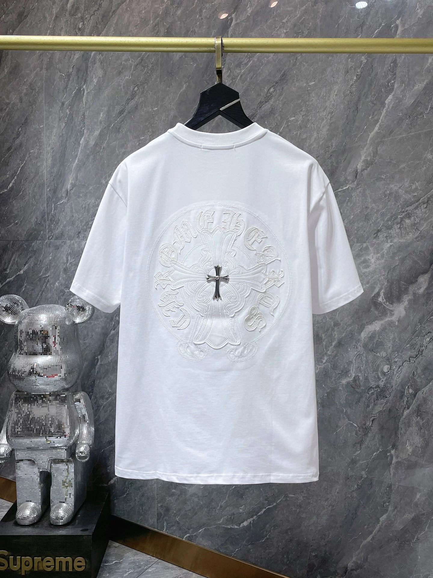 White t-shirt with a black cross design on a hanger against a marble wall.