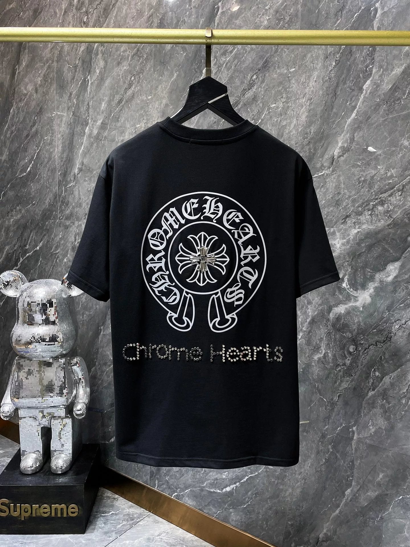 Black t-shirt with 'Chrome Hearts' logo on a hanger against a marble wall.