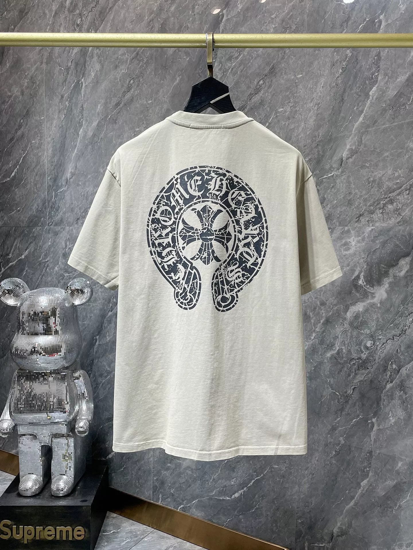 Beige t-shirt with black graphic design on a hanger against a marble wall.