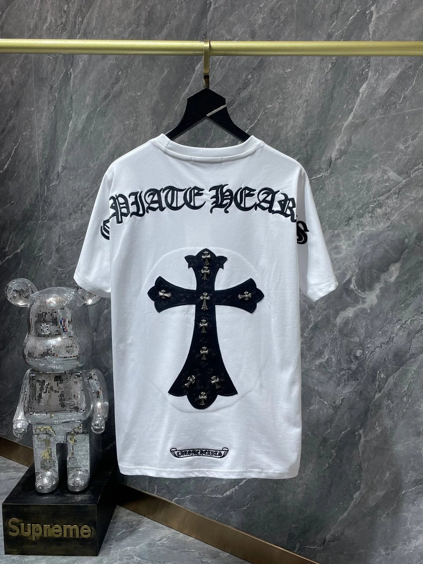 White t-shirt with black cross design on a hanger against a marble wall, next to a transparent bear figure and Supreme box.