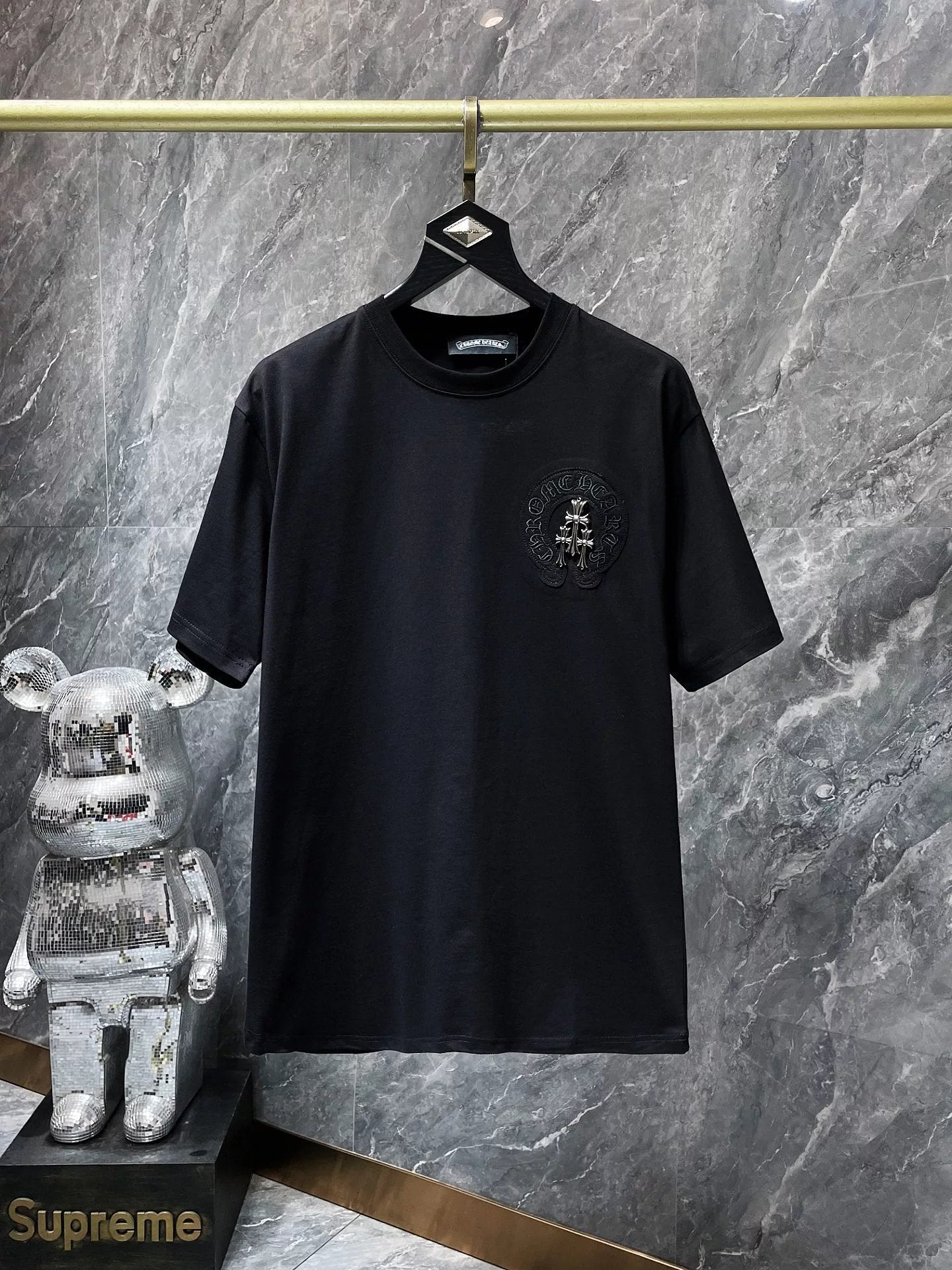 Black t-shirt with a logo on a hanger against a marble wall, with a Supreme branded object in the foreground.