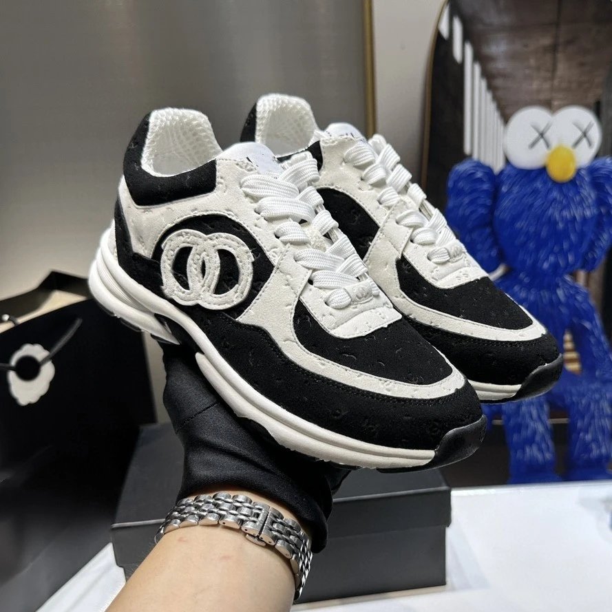 Black and white sneakers with a logo held by a gloved hand, with a blurred background.