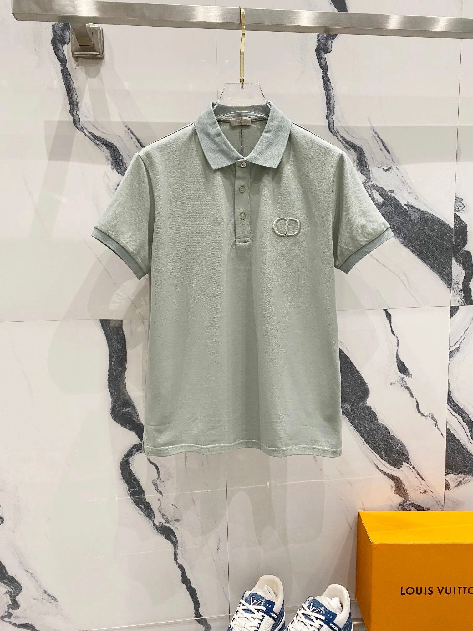 Light green polo shirt with brand logo on a hanger against a marble wall, with a Louis Vuitton box in the foreground.