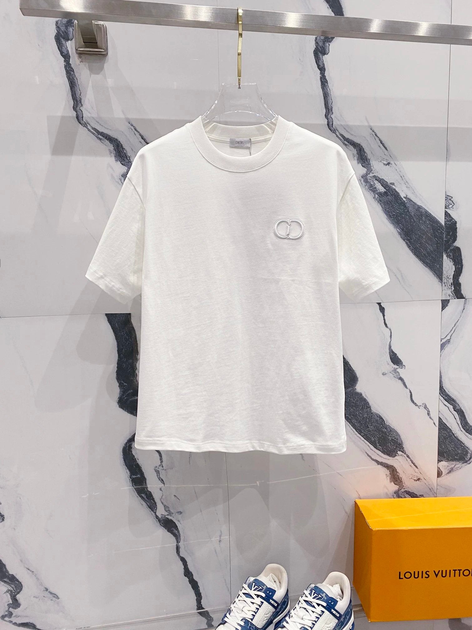 White t-shirt with a logo on a hanger against a marble wall, with a Louis Vuitton box in the foreground.