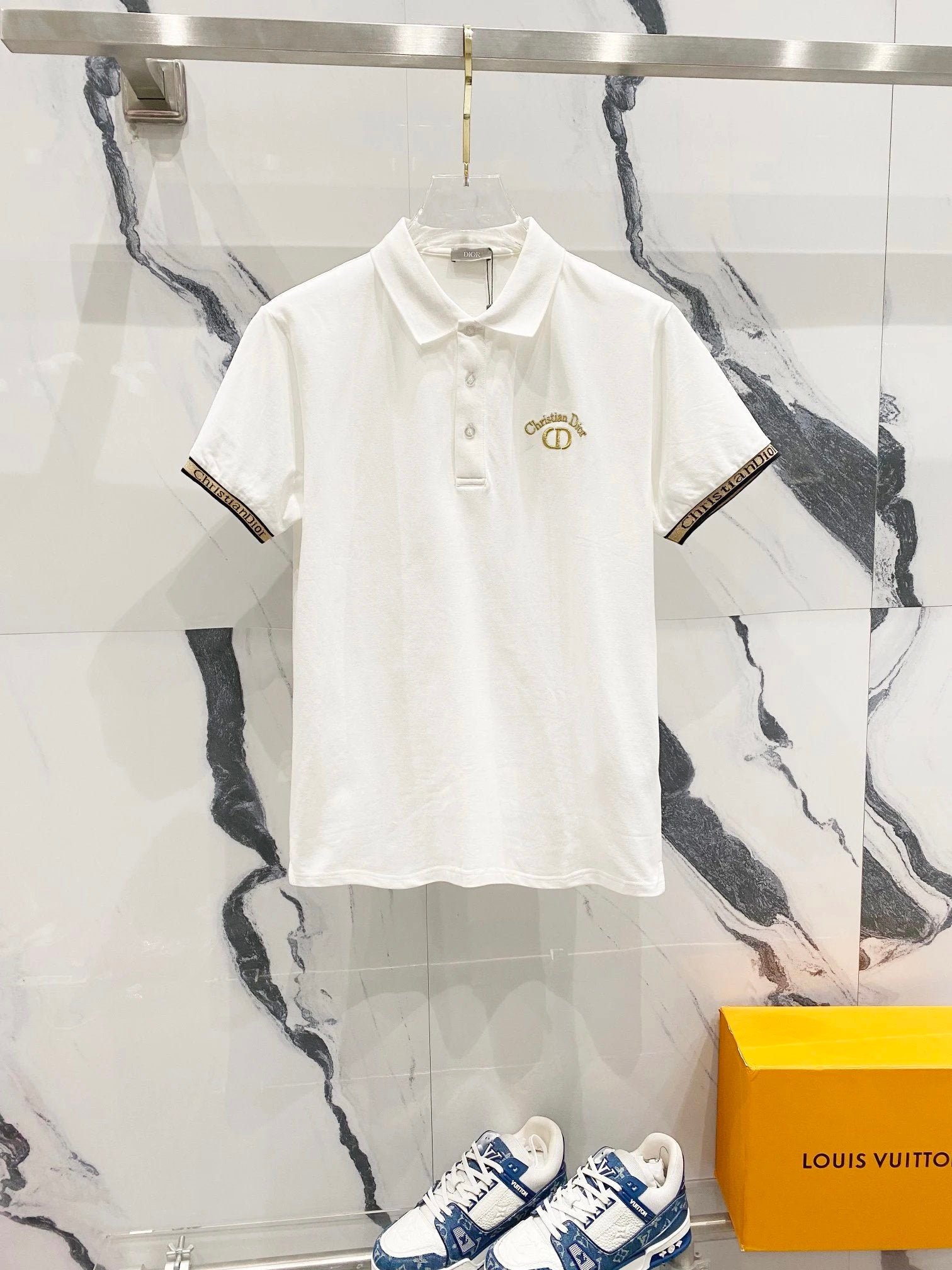 White polo shirt with gold logo on a marble background, with a Louis Vuitton sneaker and box.