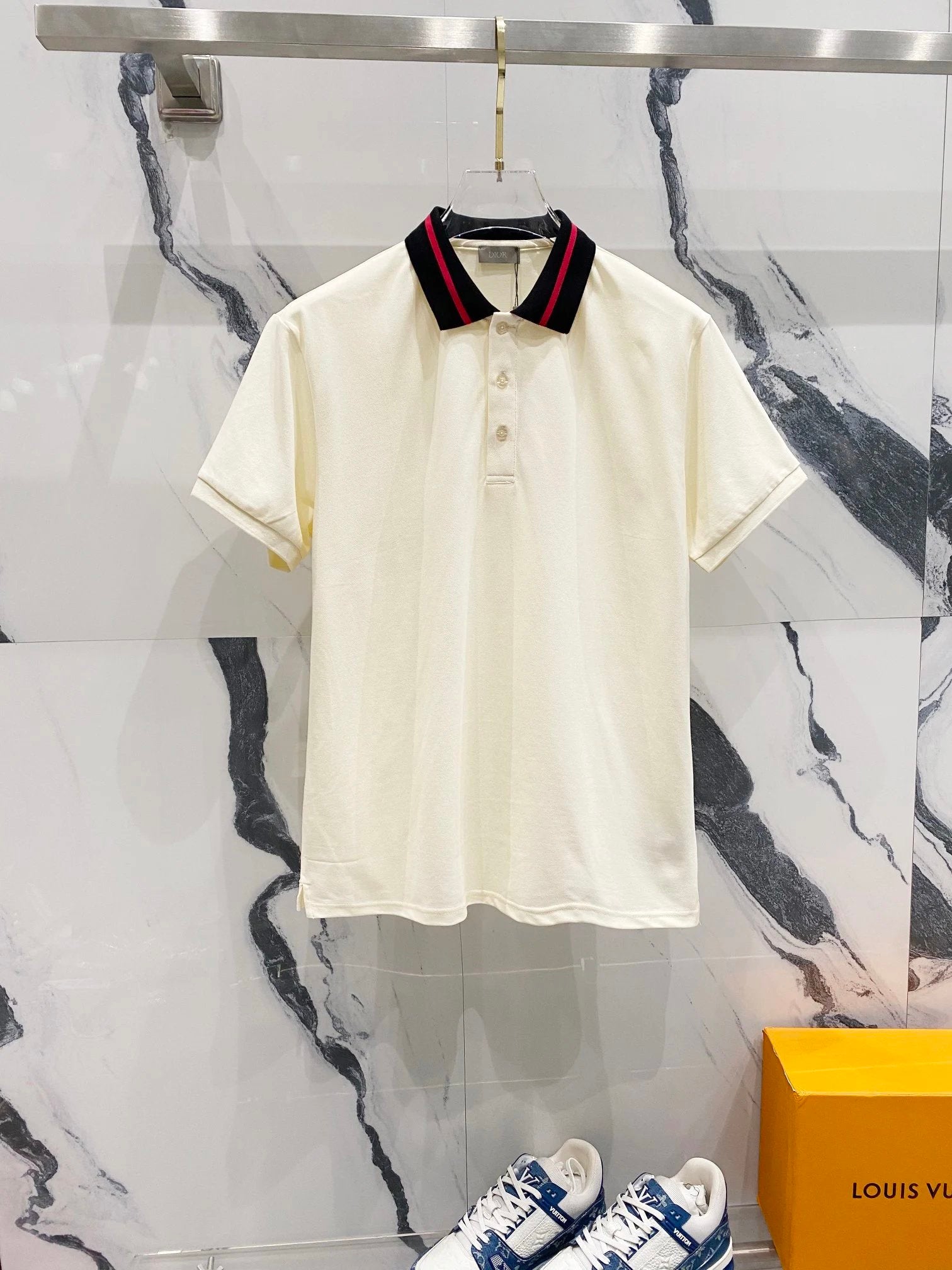 White polo shirt with red and blue collar on a hanger against a marble wall with Louis Vuitton box.